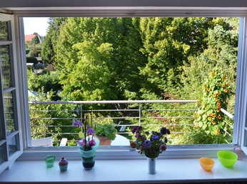 Blick aus einem offenen Fenster auf einen üppigen, grünen Garten mit Blumenarrangement im Vordergrund.