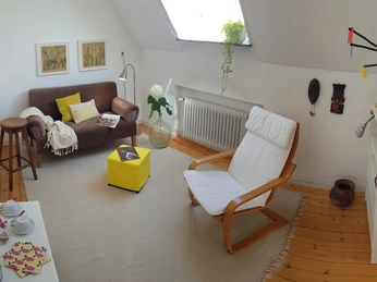 Maries B and B_Vorraum Doppelzimmer II_M Menke.jpg Heller Vorraum mit braunem Sofa, gelbem Hocker, weißem Sessel und Dekorationen an den Wänden.