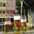 Kölschgläser stehen vor traditioneller Architektur und sprudelnder Wasserskulptur auf einem Marktplatz.