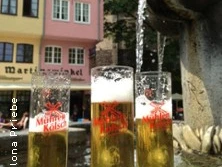 Kölschgläser stehen vor traditioneller Architektur und sprudelnder Wasserskulptur auf einem Marktplatz.