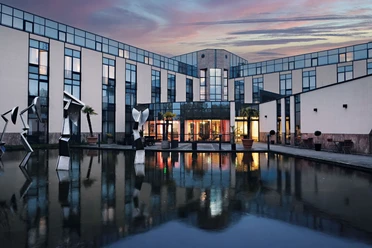 Außenansicht des Four Points Flex by Sheraton Ratingen Düsseldorf Airport Moderne Hotelfassade mit Glasflächen, Skulpturen und Wasserbecken bei Abenddämmerung in Ratingen.