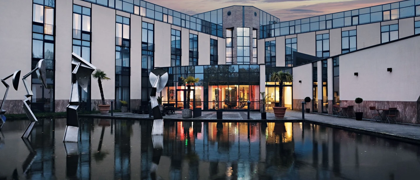 Außenansicht des Four Points Flex by Sheraton Ratingen Düsseldorf Airport Moderne Hotelfassade mit Glasflächen, Skulpturen und Wasserbecken bei Abenddämmerung in Ratingen.