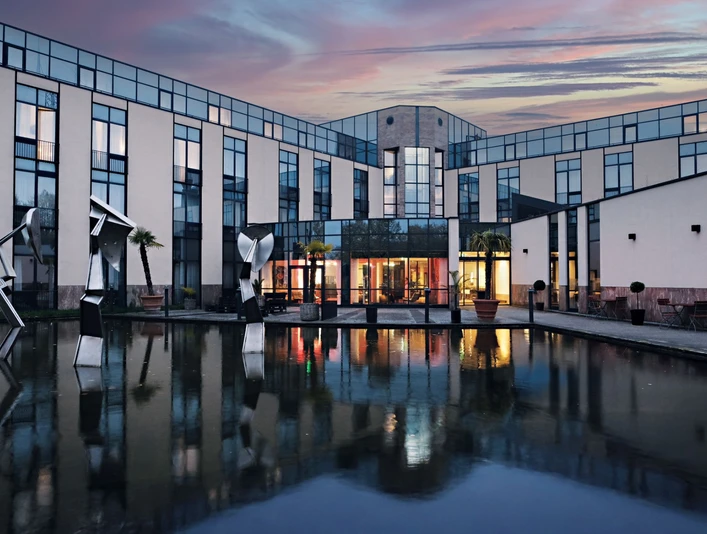 Außenansicht des Four Points Flex by Sheraton Ratingen Düsseldorf Airport Moderne Hotelfassade mit Glasflächen, Skulpturen und Wasserbecken bei Abenddämmerung in Ratingen.