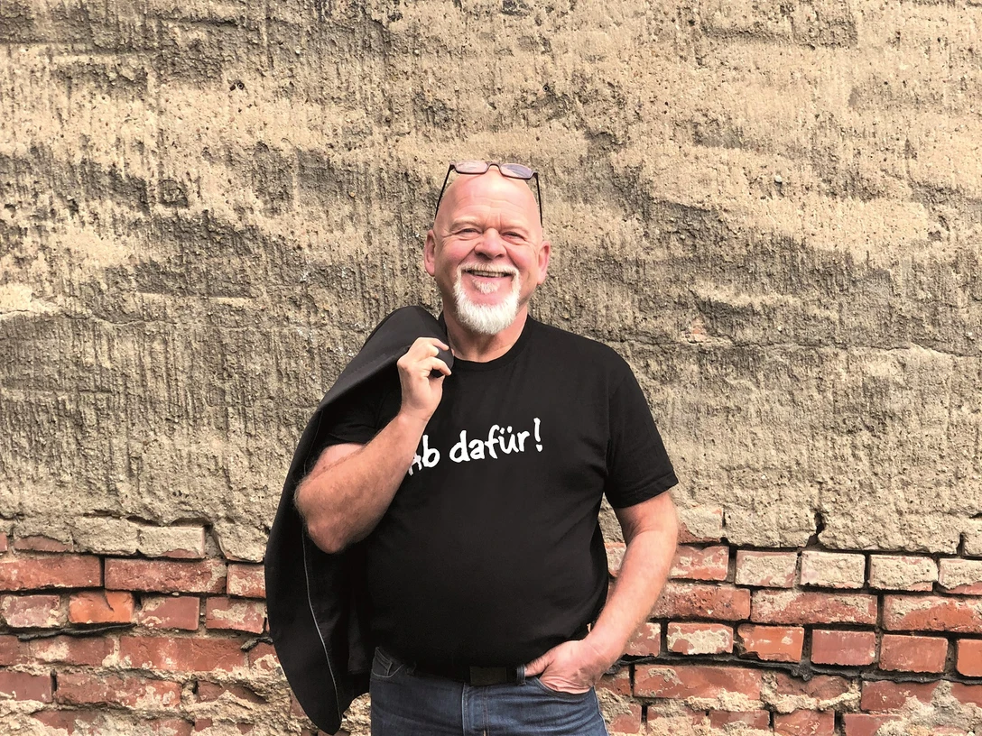 Bernd Gieseking Ein lächelnder Mann mit Bart und Brille lehnt entspannt an einer Ziegelwand, trägt Jeans und T-Shirt.