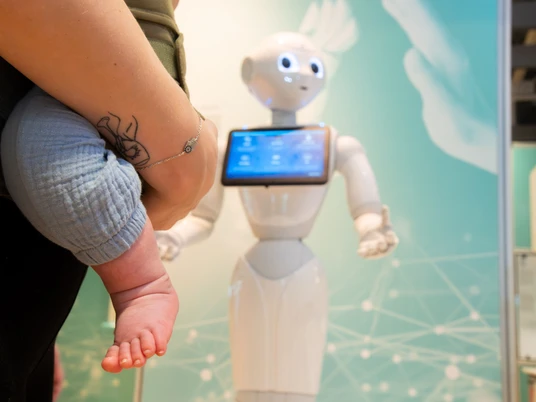 Känguru_Führung_HNF-2.jpg Mensch hält Baby auf dem Arm, während humanoider Roboter mit Bildschirm im Hintergrund steht.