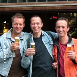 Drei Personen stehen vor einem Lokal und halten jeweils ein Glas Bier in der Hand. Sie lächeln in die Kamera.