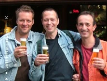 Drei Personen stehen vor einem Lokal und halten jeweils ein Glas Bier in der Hand. Sie lächeln in die Kamera.