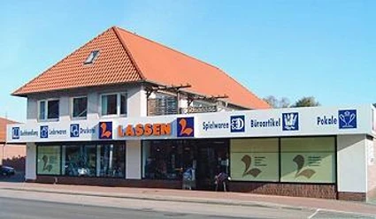 Schreibwaren Lassen.jpg Das Bild zeigt ein zweistöckiges Gebäude mit roten Dachziegeln, in dem sich ein Geschäft namens "Lassen" befindet. Große Schaufenster und eine helle Fassade zeichnen das Ladengeschäft aus. An der Fassade ist in Großbuchstaben der Name "Lassen" und Symbole für verschiedene Waren sichtbar.