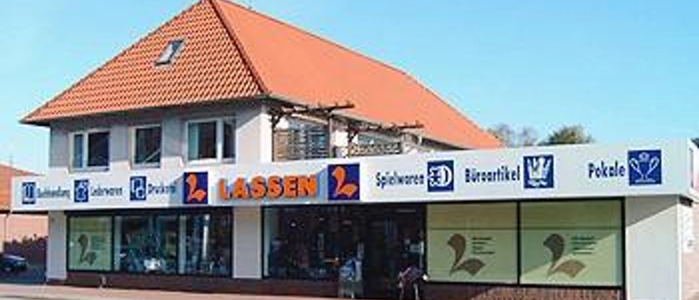Schreibwaren Lassen.jpg Das Bild zeigt ein zweistöckiges Gebäude mit roten Dachziegeln, in dem sich ein Geschäft namens "Lassen" befindet. Große Schaufenster und eine helle Fassade zeichnen das Ladengeschäft aus. An der Fassade ist in Großbuchstaben der Name "Lassen" und Symbole für verschiedene Waren sichtbar.