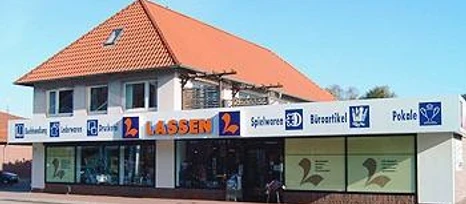 Schreibwaren Lassen.jpg Das Bild zeigt ein zweistöckiges Gebäude mit roten Dachziegeln, in dem sich ein Geschäft namens "Lassen" befindet. Große Schaufenster und eine helle Fassade zeichnen das Ladengeschäft aus. An der Fassade ist in Großbuchstaben der Name "Lassen" und Symbole für verschiedene Waren sichtbar.
