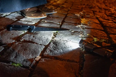 Krimitour3.jpg Auf dem Bild sind ein Taschenlampe und ein Messer auf einem gepflasterten Boden zu sehen. Eine im Dunkeln liegende Szene, die von schummrigem Licht beleuchtet wird und durch herabgefallenes Laub ergänzt wird, schafft eine mysteriöse, krimihafte Atmosphäre.