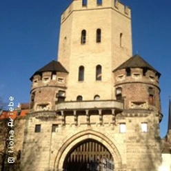 Mittelalterlicher Stadtturm mit runden Ecktürmen und Torbogen, umgeben von blauem Himmel.