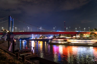 The night shot shows an illuminated bridge spanning a river. The river reflects the colorful rays of light emanating from a passing ship, while silhouettes of buildings and the gloomy evening sky can be seen in the background.
