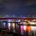 Die Nachtaufnahme zeigt eine beleuchtete Brücke, die über einen Fluss spannt. Der Fluss reflektiert die bunten Lichtstrahlen, die von einem vorbeifahrenden Schiff ausgehen, während im Hintergrund Silhouetten von Gebäuden und die düstere Abendhimmel zu sehen sind.The night shot shows an illuminated bridge spanning a river. The river reflects the colorful rays of light emanating from a passing ship, while silhouettes of buildings and the gloomy evening sky can be seen in the background.De nachtopname toont een verlichte brug over een rivier. De rivier weerspiegelt de kleurrijke lichtstralen van een passerend schip, terwijl op de achtergrond silhouetten van gebouwen en de sombere avondlucht te zien zijn.