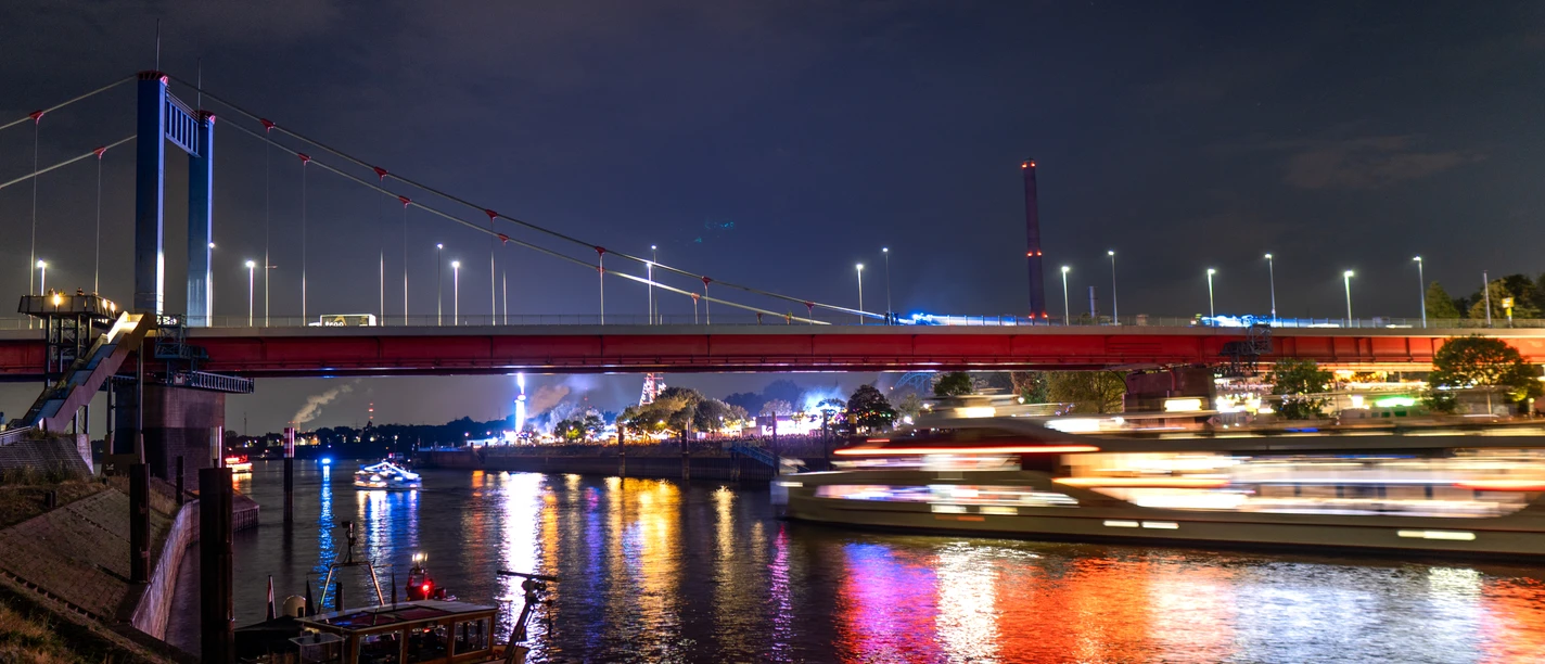 The night shot shows an illuminated bridge spanning a river. The river reflects the colorful rays of light emanating from a passing ship, while silhouettes of buildings and the gloomy evening sky can be seen in the background.