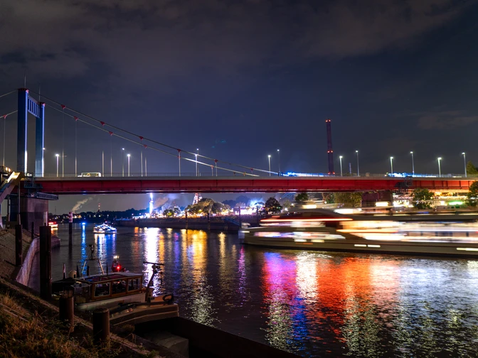 Die Nachtaufnahme zeigt eine beleuchtete Brücke, die über einen Fluss spannt. Der Fluss reflektiert die bunten Lichtstrahlen, die von einem vorbeifahrenden Schiff ausgehen, während im Hintergrund Silhouetten von Gebäuden und die düstere Abendhimmel zu sehen sind.The night shot shows an illuminated bridge spanning a river. The river reflects the colorful rays of light emanating from a passing ship, while silhouettes of buildings and the gloomy evening sky can be seen in the background.De nachtopname toont een verlichte brug over een rivier. De rivier weerspiegelt de kleurrijke lichtstralen van een passerend schip, terwijl op de achtergrond silhouetten van gebouwen en de sombere avondlucht te zien zijn.