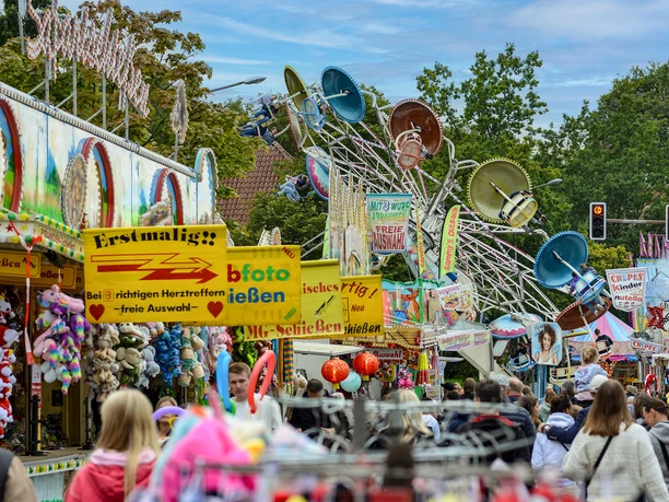 Zwiebelmarkt für Destination One 7.jpg Menschen schlendern über einen belebten Jahrmarkt mit bunten Ständen und einem Fahrgeschäft.