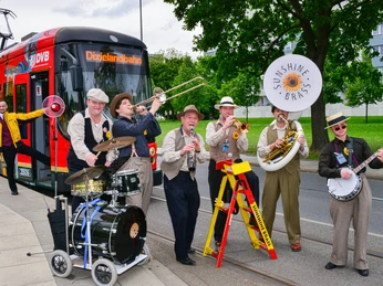 Dixielandfestival-Dixietram-Jazzrundfahrt-1-©-Foto-Hendrik-Meyer.jpg Eine Jazzband spielt vor einer roten Straßenbahn auf einem Gehweg.