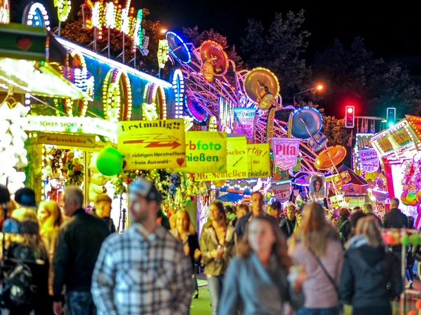 Zwiebelmarkt für Destination One 2.jpg Menschen genießen lebhaftes Treiben auf einem beleuchteten Jahrmarkt, umgeben von bunten Fahrgeschäften.