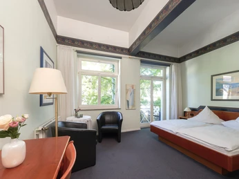 Zimmerbeispiel DZ Helles Doppelzimmer mit großen Fenstern, Balkon, Doppelbett und gemütlicher Sitzgelegenheit.