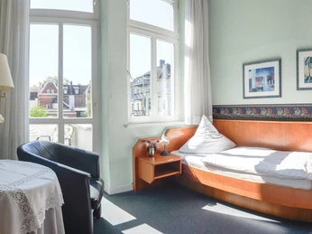 Zimmerbeispiel EZ Helles Einzelzimmer mit großem Fenster, einem Einzelbett, Ledersessel und Tisch mit Blumenvase.