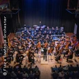 Das Bild zeigt ein Orchester auf einer Bühne, das von einem Dirigenten geleitet wird, umgeben von Zuschauenden.