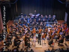 Das Bild zeigt ein Orchester auf einer Bühne, das von einem Dirigenten geleitet wird, umgeben von Zuschauenden.