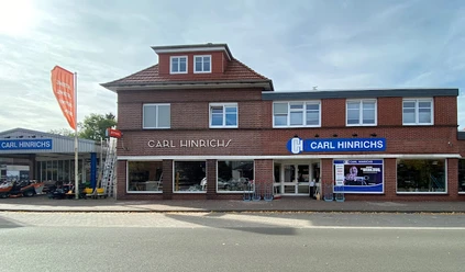 Carl Hinrichs.jpg Das Bild zeigt ein zweistöckiges, rotes Backsteingebäude mit dem Schriftzug "Carl Hinrichs" über den Schaufenstern. Rechts daneben ein modernes Gebäude mit einer großen Glasfront und einem blauen Logo. Vor dem Gebäude verläuft eine Straße. Im Hintergrund sind ein Fahnenmast und ein Baum zu erkennen.