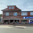 Carl Hinrichs.jpg Das Bild zeigt ein zweistöckiges, rotes Backsteingebäude mit dem Schriftzug "Carl Hinrichs" über den Schaufenstern. Rechts daneben ein modernes Gebäude mit einer großen Glasfront und einem blauen Logo. Vor dem Gebäude verläuft eine Straße. Im Hintergrund sind ein Fahnenmast und ein Baum zu erkennen.