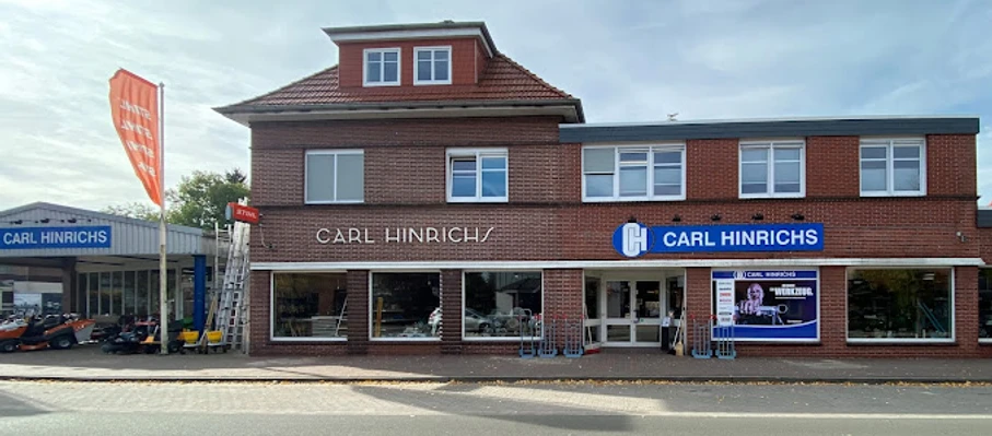 Carl Hinrichs.jpg Das Bild zeigt ein zweistöckiges, rotes Backsteingebäude mit dem Schriftzug "Carl Hinrichs" über den Schaufenstern. Rechts daneben ein modernes Gebäude mit einer großen Glasfront und einem blauen Logo. Vor dem Gebäude verläuft eine Straße. Im Hintergrund sind ein Fahnenmast und ein Baum zu erkennen.