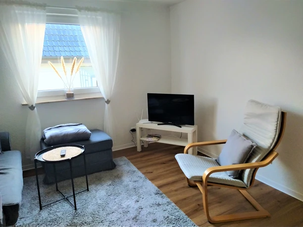 Wohnbereich mit grauem Sofa, Sessel, flachem Fernseher, hellem Holzboden und Tageslicht von links.