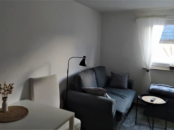 Herford_FeWo Suedstadt__Wohnbereich_Matuschat (1).jpg Wohnbereich mit grauem Sofa, soften Kissen, Leselampe, kleinem Tisch und Fenster mit weißem Vorhang.