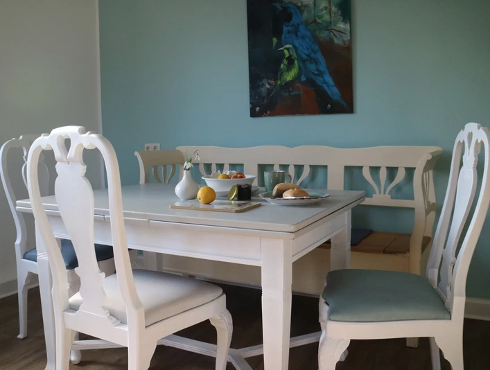 Weißer Esstisch mit vier Stühlen und blauem Bild an der Wand, gedeckt mit Obst und Kerzen.