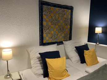 Zwei Betten mit gelben und weißen Kissen, Leselampen und einem farbenfrohen Wandteppich darüber.