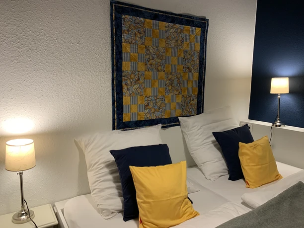 Herford_Apartment am Stiftberg_Schlafkoje_Niestrath (8).jpg Zwei Betten mit gelben und weißen Kissen, Leselampen und einem farbenfrohen Wandteppich darüber.
