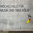 Beethoven Banana by Thomas Baumgärtel Das Bild zeigt ein Straßenkunstwerk von Thomas Baumgärtel an der Fassade der Hochschule für Musik und Tanz Köln. Es stellt eine Banane dar, die in einem stilisierten Porträt Ludwig van Beethoven ähnelt. Die Banane ist in leuchtendem Gelb gemalt, und ihre Oberseite ist so gestaltet, dass sie Beethovens charakteristisches Haar und Gesichtszüge hat.The picture shows a work of street art by Thomas Baumgärtel on the façade of the Cologne University of Music and Dance. It depicts a banana that resembles Ludwig van Beethoven in a stylized portrait. The banana is painted in bright yellow and its upper side is designed to have Beethoven's characteristic hair and facial features.