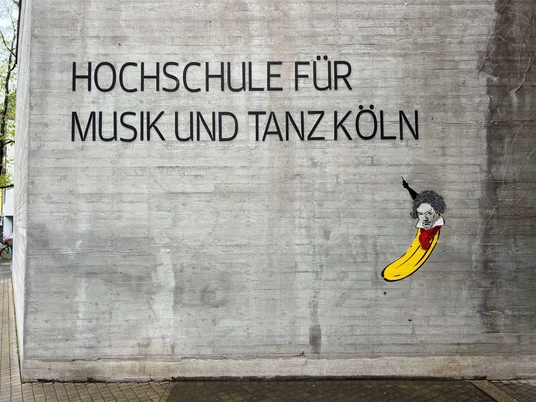 Beethoven Banane by Thomas Baumgärtel Das Bild zeigt ein Straßenkunstwerk von Thomas Baumgärtel an der Fassade der Hochschule für Musik und Tanz Köln. Es stellt eine Banane dar, die in einem stilisierten Porträt Ludwig van Beethoven ähnelt. Die Banane ist in leuchtendem Gelb gemalt, und ihre Oberseite ist so gestaltet, dass sie Beethovens charakteristisches Haar und Gesichtszüge hat.The picture shows a work of street art by Thomas Baumgärtel on the façade of the Cologne University of Music and Dance. It depicts a banana that resembles Ludwig van Beethoven in a stylized portrait. The banana is painted in bright yellow and its upper side is designed to have Beethoven's characteristic hair and facial features.