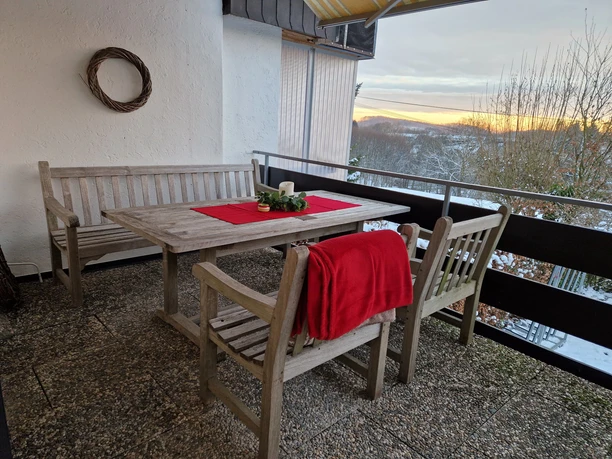 Terrasse Winterliche Terrasse mit Holzmöbeln, Tischdeko und Blick auf verschneite Bäume bei Sonnenuntergang.