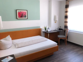 Ein helles Hotelzimmer mit Einzelbett, Schreibtisch, Stuhl und Lampe vor einem Fenster mit Vorhängen.