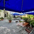 Herford-HotelPfennigskrug-Biergarten-H Althoff.jpg Biergarten im Freien mit Holztischen und blauen Sonnenschirmen, umgeben von grünen Pflanzen.
