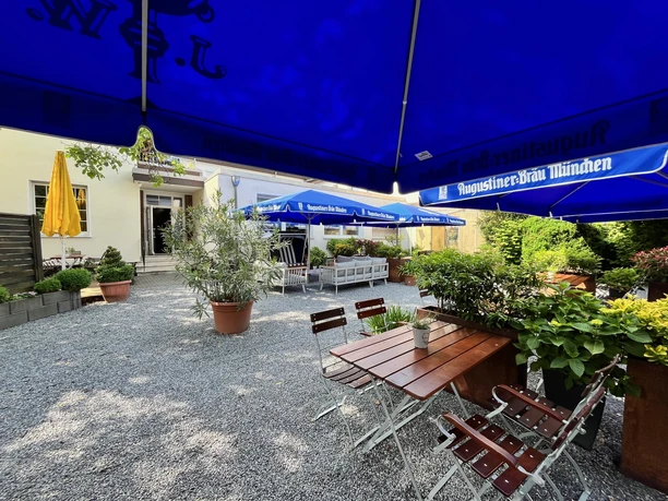 Herford-HotelPfennigskrug-Biergarten-H Althoff.jpg Biergarten im Freien mit Holztischen und blauen Sonnenschirmen, umgeben von grünen Pflanzen.