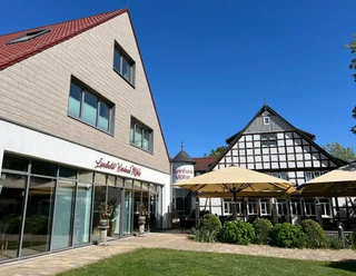 Weinhaus Front Zwei Gebäude mit roten Dächern und Fachwerk, umgeben von Pflanzen und Sonnenschirmen im Hof.