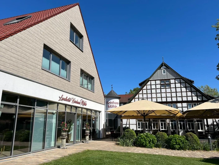 Weinhaus Front Zwei Gebäude mit roten Dächern und Fachwerk, umgeben von Pflanzen und Sonnenschirmen im Hof.