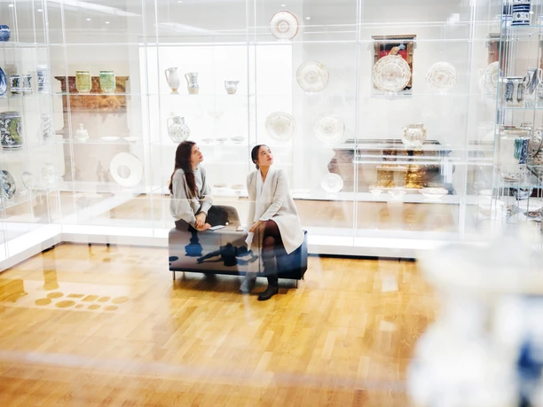 GRASSI Museum für Angewandte Kunst - Von Jugendstil bis Gegenwart Zwei Frauen Sitzen inmitten gläserner Vitrinen mit Ausstellungsstücken wie Tellern und Kannen im GRASSI Museum für Angewandte Kunst
