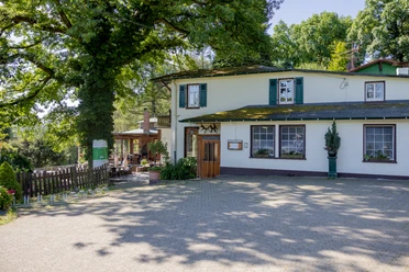 Zweistöckiges weißes Gasthaus im Grünen, umgeben von Bäumen. Elegante Terrasse im Hintergrund.
