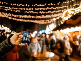 Weihnachtsdorf in Einbeck im Lichterglanz Hand, die eine Tasse Glühwein hält im Fokus und das beleuchtete Einbecker Weihnachtsdorf dahinter.Hand holding a cup of mulled wine in focus and the illuminated Einbeck Christmas village behind it.En hånd med en kop gløgg i fokus og den oplyste Einbeck-juleby bagved.Hand die een kop glühwein vasthoudt in focus en het verlichte Einbeck kerstdorp erachter.