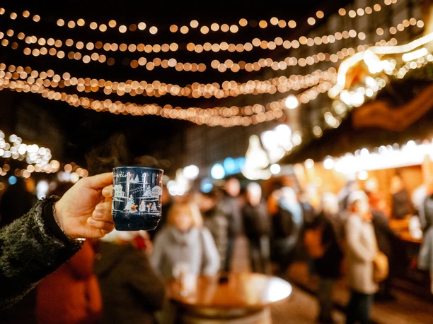 Weihnachtsdorf in Einbeck im Lichterglanz Hand, die eine Tasse Glühwein hält im Fokus und das beleuchtete Einbecker Weihnachtsdorf dahinter.