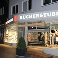 Lübbecker Bücherstube.JPG Leuchtende Buchhandlung mit offener Glastür, Schaufenstern voller Bücher in der Abenddämmerung.
