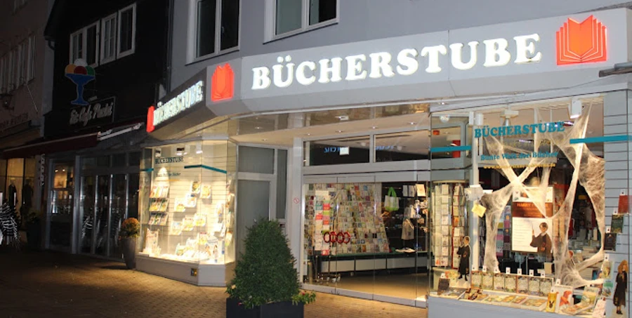 Lübbecker Bücherstube.JPG Leuchtende Buchhandlung mit offener Glastür, Schaufenstern voller Bücher in der Abenddämmerung.
