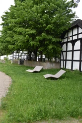 Zwei Holzliegen stehen auf einer Wiese unter einem großen Baum neben einem Fachwerkhaus.